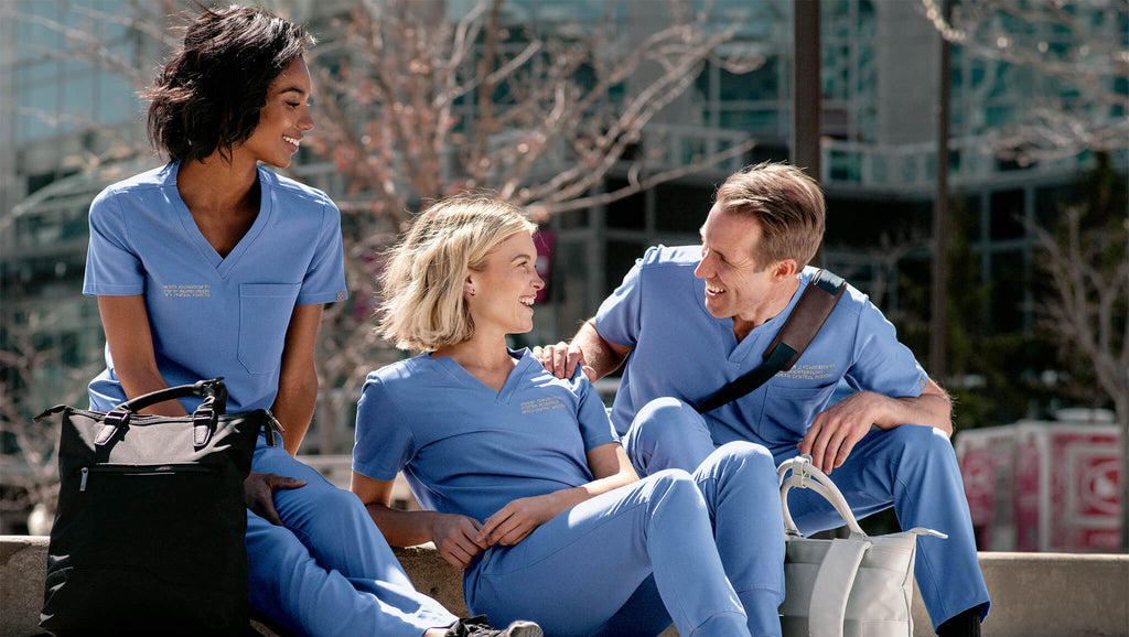 Two women and one man are sitting on the steps enjoying each other's company. They are all wearing MedTailor outfits consisting of a scrub top and pants in Sky Blue color fabric.