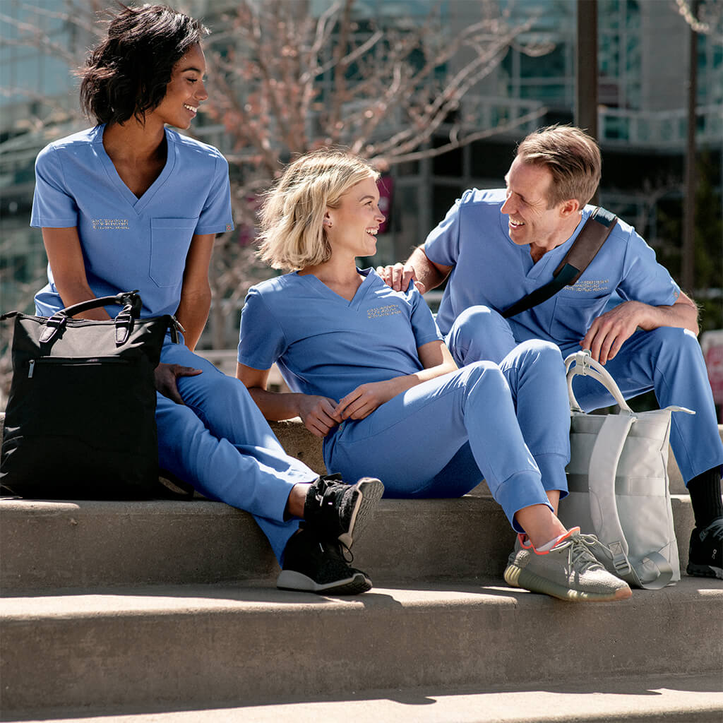 Two women and one man are sitting on the steps enjoying each other's company. They are all wearing MedTailor outfits consisting of a scrub top and pants in Sky Blue color fabric.