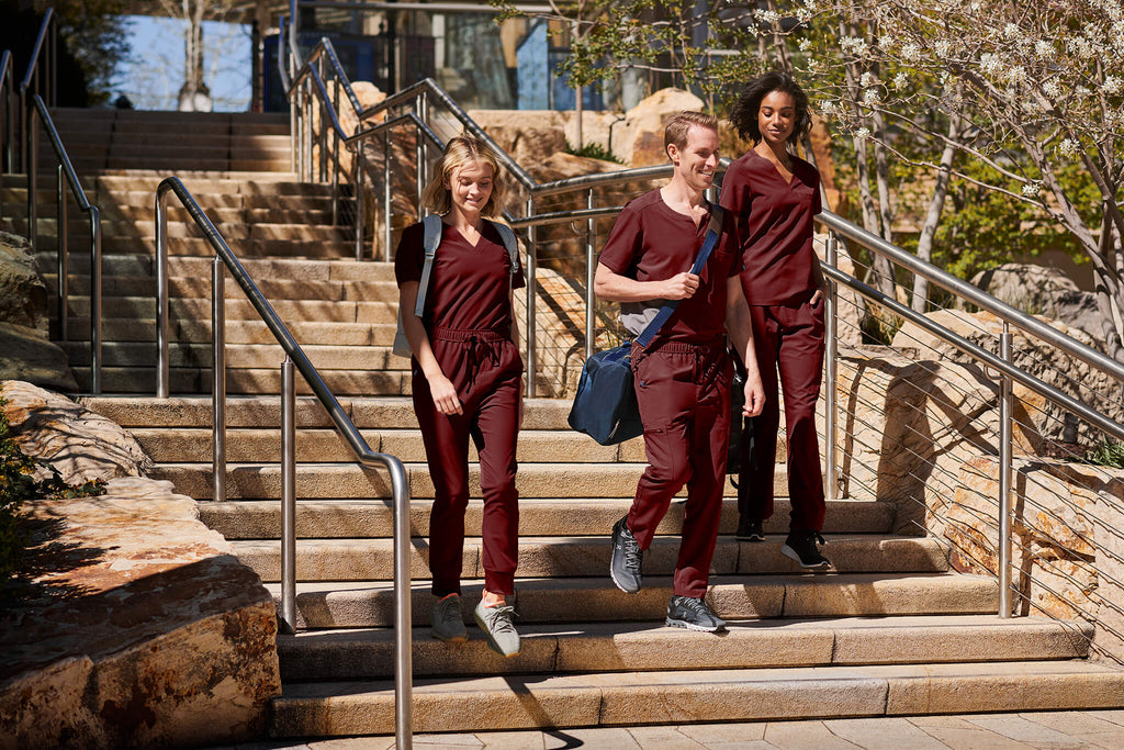 3 friends are wearing MedTailor scrubs in Merlot Red. They are walking down a flight of stairs together.