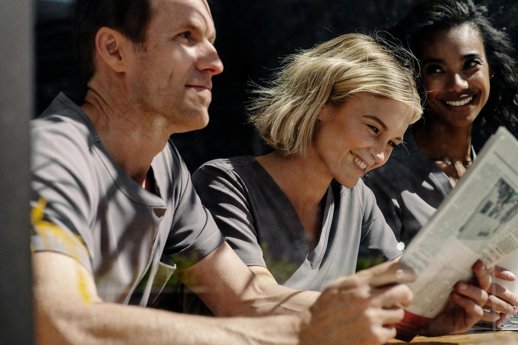 3 friends wearing MedTailor scrubs in Platinum Gray color fabric. They are sitting at a coffee shop counter facing the street window, 