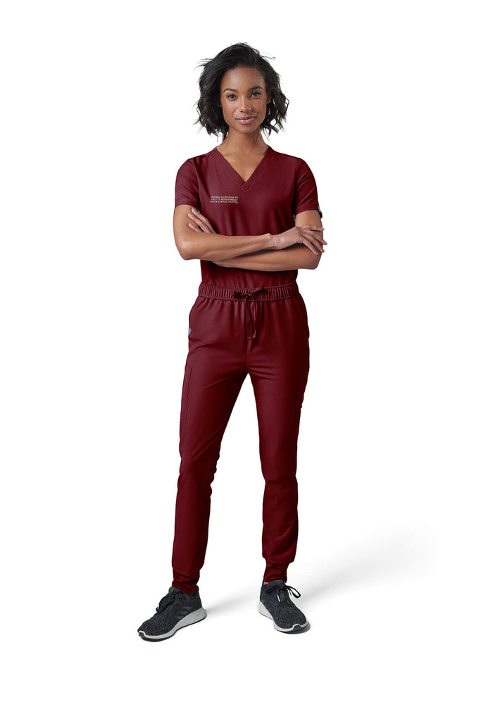 A woman wearing MedTailor women's scrub top in Merlot Red color fabric. She is standing and crossing her arms.
